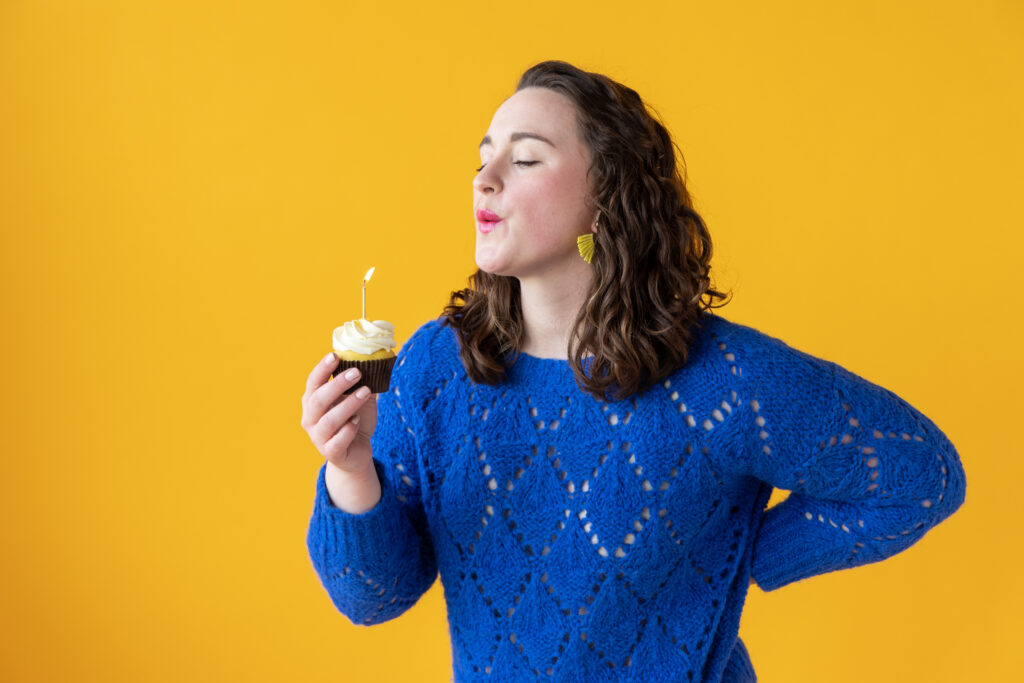 Woman blowing out the candle on a cupcake, against a bright yellow backdrop.
