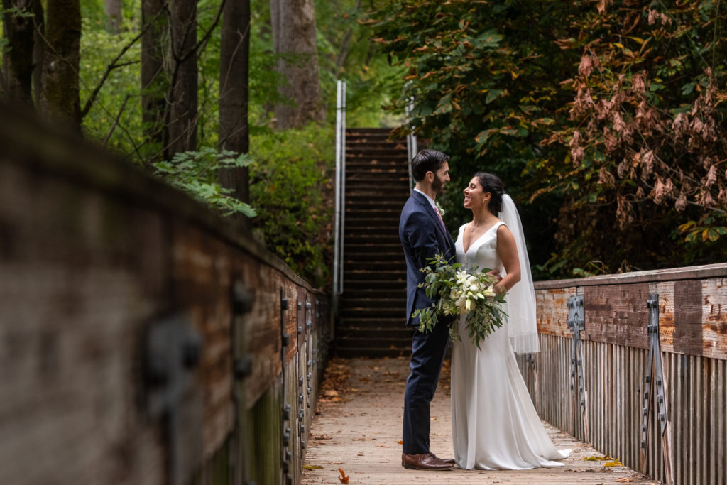 Bride and groom standing on wooden bridge, looking at each other.
