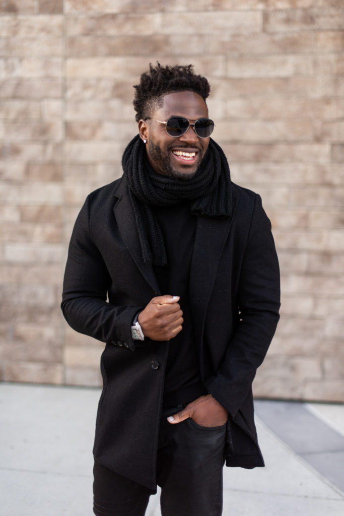 Men's portrait, subject wearing black sunglasses, black scarf, and all-black outfit, smiling.