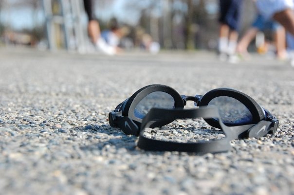 photo of black swim goggles on the pavement from a low perspective, showing a shallow depth of field, with people's legs and feet blurred in the background.