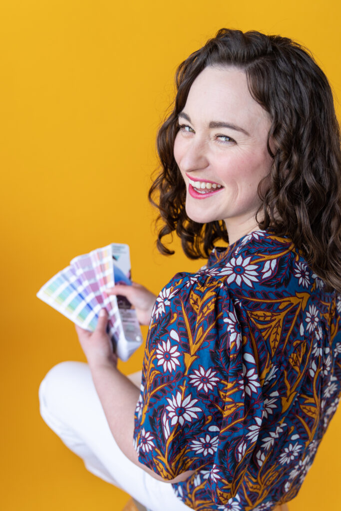 Woman holding color swatches, sitting against a bright yellow backdrop.