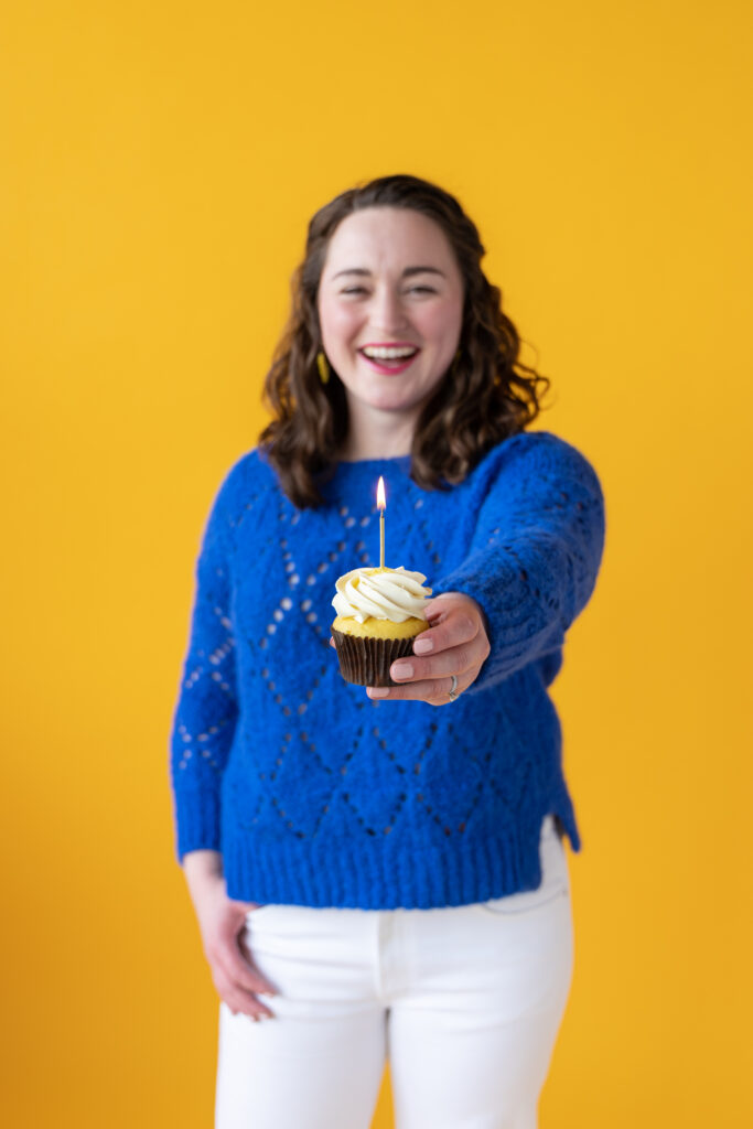 Woman holding a cupcake with candle towards the camera, against a bright yellow backdrop.